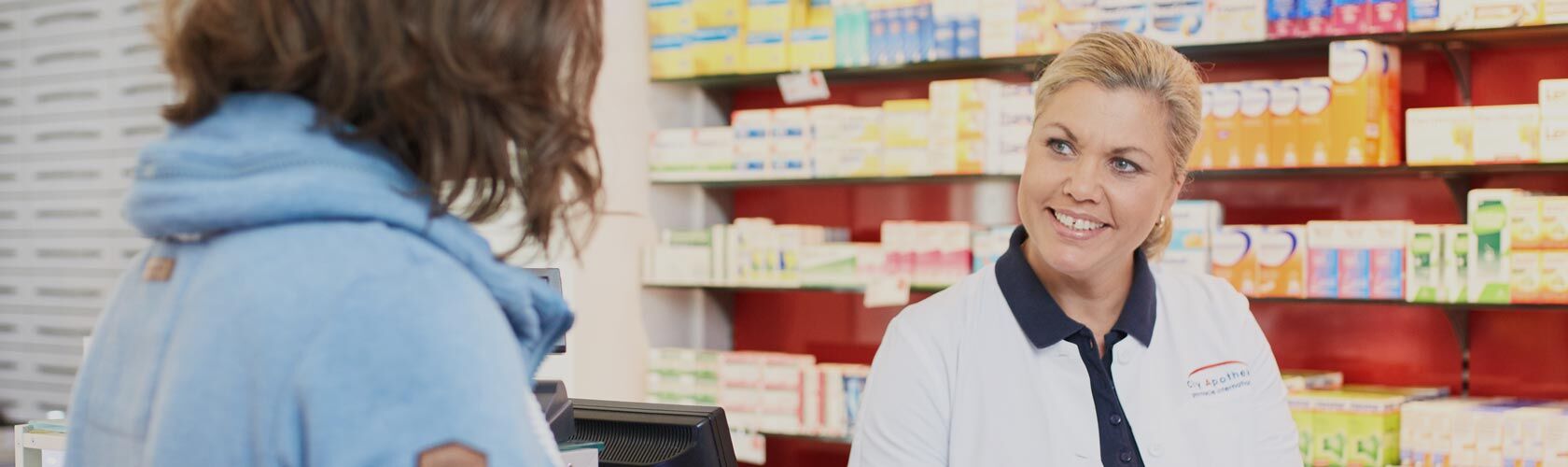 Eine freundliche Mitarbeiterin berät eine Kundin vor einem Regal mit Medikamenten in der Apotheke.