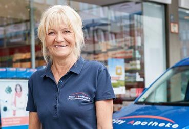 Eine lächelnde Frau in einem "City Apotheke"-Shirt steht vor einem blauen Lieferfahrzeug.